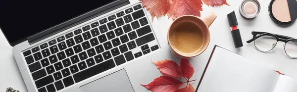 Plan panoramique de l'ordinateur portable près de tasse à café, ordinateur portable, verres, cosmétiques et feuilles rouges de raisins sauvages sur table blanche — Photo de stock