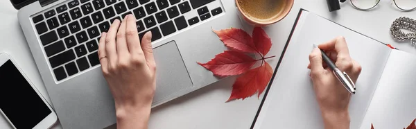 Vue recadrée de la femme à l'aide d'un ordinateur portable et tenant un stylo près du portable, smartphone, tasse à café et feuille rouge de raisins sauvages sur table blanche, prise de vue panoramique — Photo de stock