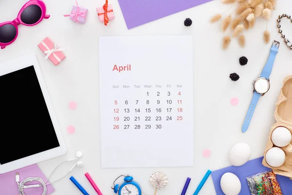 Página del calendario de abril, tableta digital, gafas de sol, aspersiones de pastelería, huevos de pollo, mora, espiguillas, reloj de pulsera, reloj despertador de juguete, auriculares aislados en blanco - foto de stock