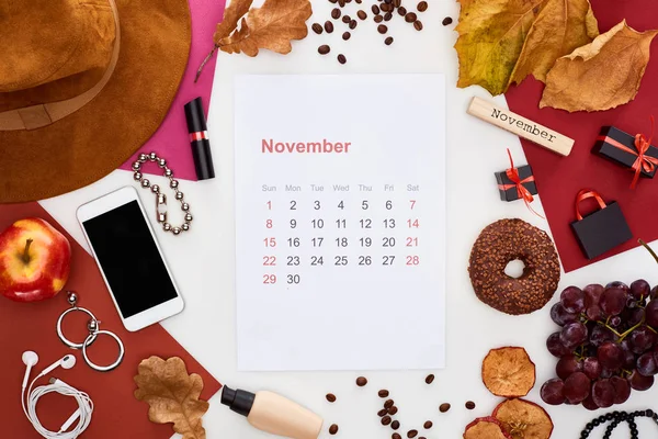 November calendar page, hat, smartphone, fruits, cosmetics, dry leaves, donut, multicolored papers, wooden block with november inscription isolated on white — Stock Photo