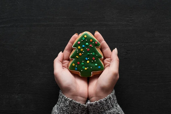 Vista ritagliata della donna che tiene il biscotto dell'albero di Natale in mano sul tavolo di legno nero — Foto stock