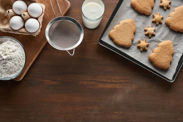 Vista superior de galletas de Navidad en la bandeja del horno cerca de los ingredientes en la mesa de madera - foto de stock