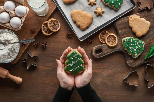 Cropped view of woman holding Christmas tree cookie near ingredients and spices on wooden table — Stock Photo