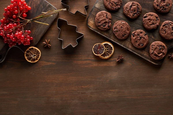 Vista superior de biscoitos de chocolate perto de moldes de massa de Natal, viburnum e especiarias na mesa de madeira — Fotografia de Stock