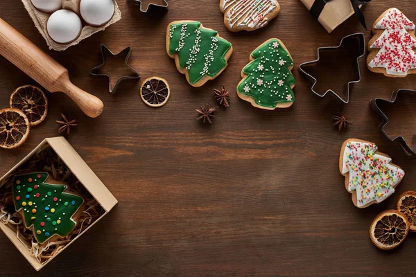 Vista superior de las galletas del árbol de Navidad, caja de regalo, moldes de masa, huevos y rodillo en la mesa de madera - foto de stock