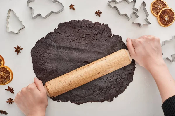 Vista recortada de la mujer sosteniendo el rodillo en la masa cruda para galletas de Navidad de chocolate sobre fondo blanco cerca de anís, moldes de masa y cítricos secos - foto de stock