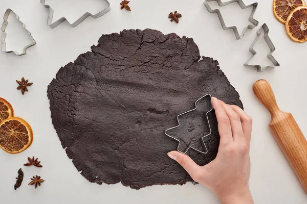 Vista recortada de la mujer sosteniendo molde de masa de árbol de Navidad en masa cruda sobre fondo blanco cerca de anís, moldes de masa, rodillo y cítricos secos - foto de stock