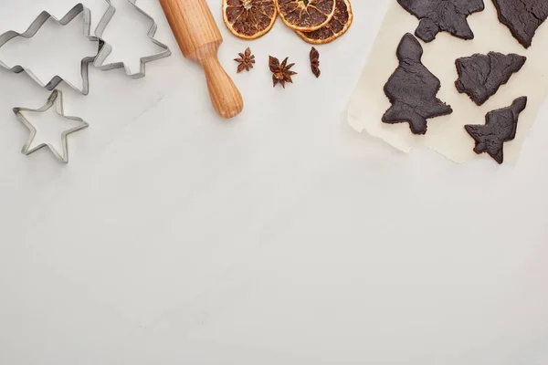 Vista superior de la masa cruda para galletas de Navidad de chocolate con rodillo sobre fondo blanco cerca de anís, moldes de masa y cítricos secos - foto de stock