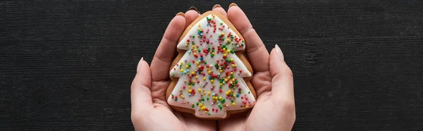 Cropped view of woman holding Christmas tree cookie in hands on black wooden table, panoramic shot — Stock Photo