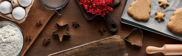 Top view of baked Christmas cookies near ingredients, dough molds and viburnum on wooden table, panoramic shot — Stock Photo