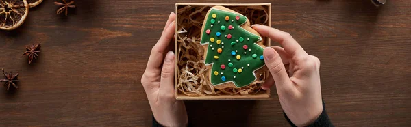 Vista recortada de la mujer poniendo galletas del árbol de Navidad en la caja de regalo en la mesa de madera, tiro panorámico - foto de stock