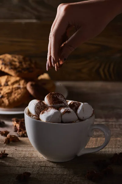 Cropped view of woman sprinkling cacao powder on marshmallow in cup on wooden table with anise — Stock Photo