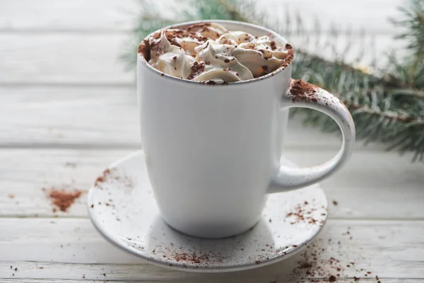 Weihnachtskakao mit Kakaopulver und Schlagsahne im Becher auf weißem Holztisch neben Tannenzweig — Stockfoto