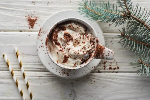 Weihnachtskakao mit Kakaopulver und Schlagsahne im Becher auf weißem Holztisch neben Tannenzweig — Stockfoto