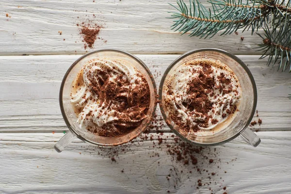 Top view of Christmas cacao with whipped cream and cacao powder in mugs on white wooden table with fir branch — Stock Photo