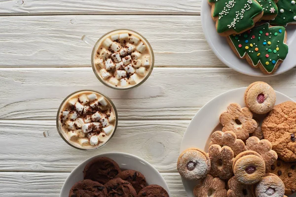 Vista dall'alto del cacao con marshmallow e cacao in polvere in tazze vicino ai biscotti natalizi su tavolo di legno bianco — Foto stock