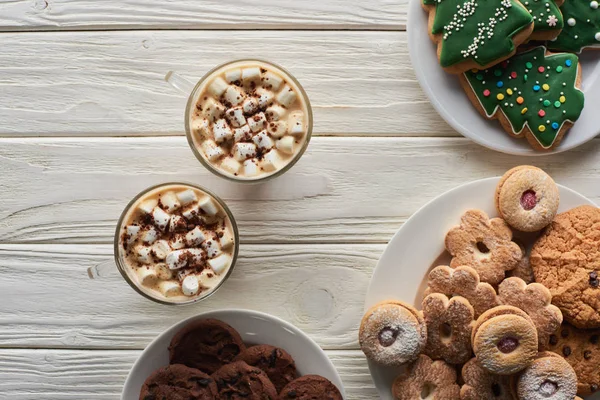 Top view of cacao with marshmallow and cacao powder in mugs near Christmas cookies on white wooden table — Stock Photo