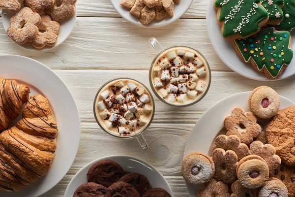 Vista superior de cacau com marshmallow e cacau em pó em canecas sobre mesa de madeira branca com biscoitos de Natal e croissants — Fotografia de Stock