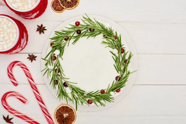 Vista superior de bastones de caramelo, dos tazas de cacao y pastel de Navidad con glaseado en la mesa de madera blanca - foto de stock