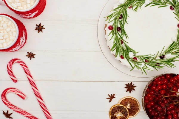 Vista dall'alto di torta di Natale, bacche di viburno, bastoncini di zucchero e due tazze di cacao con marshmallow sul tavolo di legno bianco — Foto stock