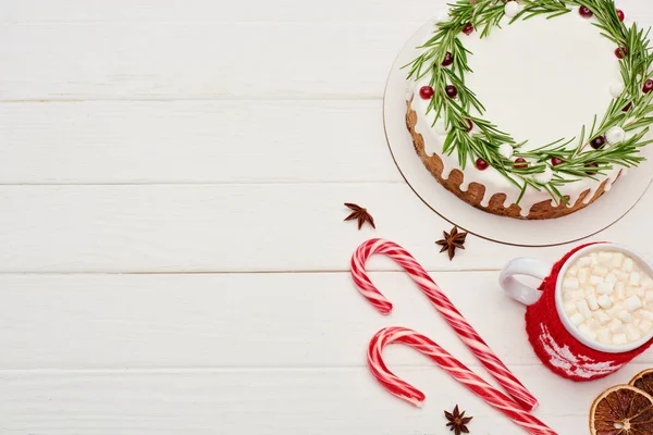 Vista superior de pastel de Navidad con glaseado en mesa de madera blanca con bastones de caramelo y taza de cacao con malvaviscos - foto de stock