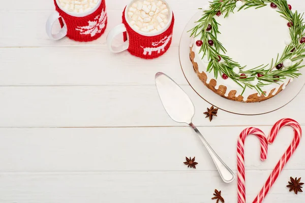 Vista superior del pastel de Navidad, bastones de caramelo y dos tazas de cacao con malvaviscos en la mesa de madera blanca - foto de stock