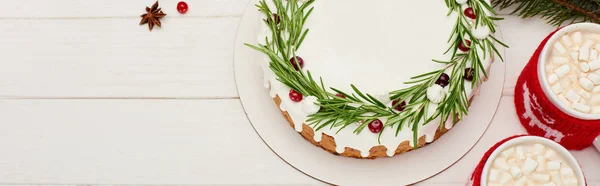 Vista superior del pastel de Navidad con glaseado, romero y arándanos sobre una mesa de madera blanca con tazas de cacao - foto de stock