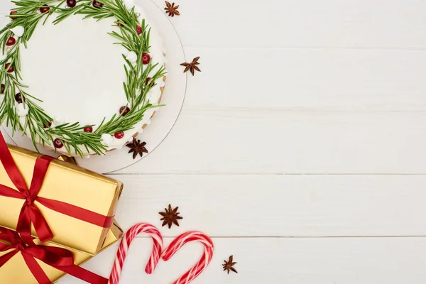 Vista superior de pastel de Navidad, semillas de estrella de anís, bastones de caramelo y cajas de regalo en mesa de madera blanca - foto de stock