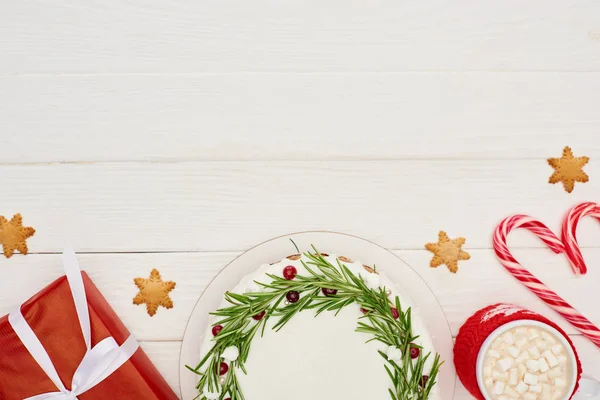 Вид зверху на різдвяний пиріг, червоний подарунок, цукерки та чашку какао з зефіром на білому дерев'яному столі — стокове фото