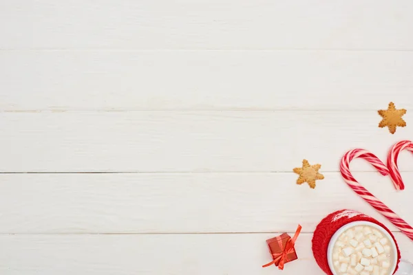 Vista superior de bastones de caramelo de Navidad, pequeño regalo, galletas y taza de cacao con malvaviscos en la mesa de madera blanca - foto de stock