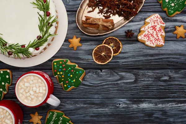 Top view of christmas pie, two cups of cocoa with marshmallows, spices and christmas tree cookies on dark wooden table — Stock Photo
