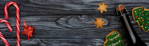 Vista superior de pequeños regalos, bastones de caramelo y galletas de árbol de Navidad y botella de champán en la mesa de madera oscura - foto de stock