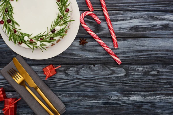 Vista superior del pastel de Navidad con romero y arándanos, bastones de caramelo, tenedor y cuchillo en la servilleta en la mesa de madera oscura — Stock Photo