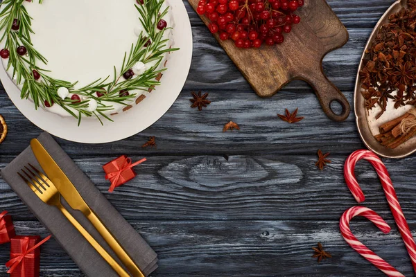 Vista superior del pastel de Navidad con bayas de romero y viburnum, regalos, tenedor y cuchillo en la servilleta en la mesa de madera oscura - foto de stock