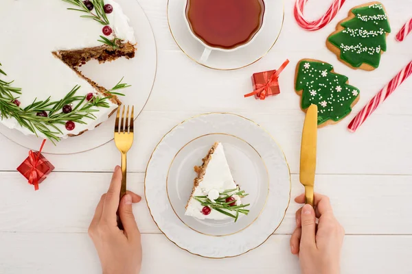 Vista ritagliata della ragazza che tiene forchetta e coltello per mangiare torta di Natale e biscotti con tè sul tavolo di legno bianco — Stock Photo