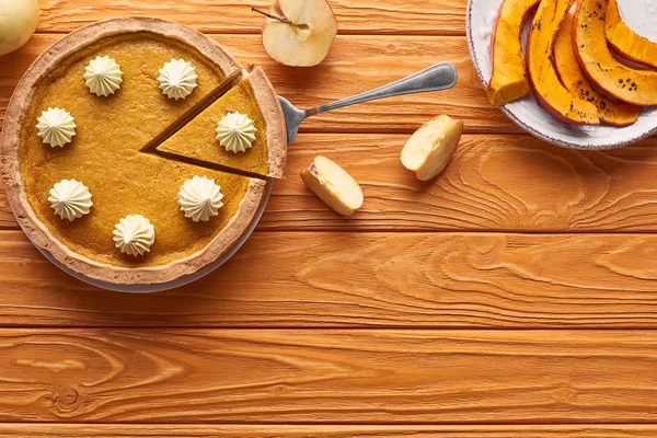 Delicioso pastel de calabaza con crema batida cerca de manzana cortada, y calabaza al horno en rodajas en la mesa de madera naranja - foto de stock
