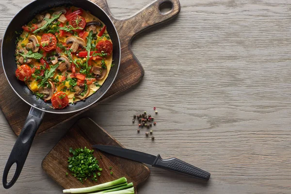 Top view of omelet in frying pan with ingredients and knife on wooden table — Stock Photo