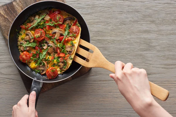 Top view of woman cooking omelet with mushrooms, tomatoes and greens on frying pan with wooden shovel — Stock Photo