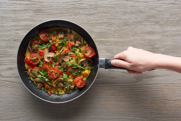 Cropped view of woman holding frying pan with omelet with mushrooms, tomatoes and greens — Stock Photo