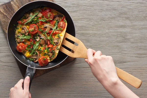 Cropped view of woman cooking omelet with mushrooms, tomatoes and greens on frying pan with wooden shovel — Stock Photo