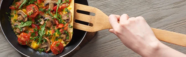 Cropped view of woman cooking tasty omelet with tomatoes and greens on frying pan with wooden shovel — Stock Photo