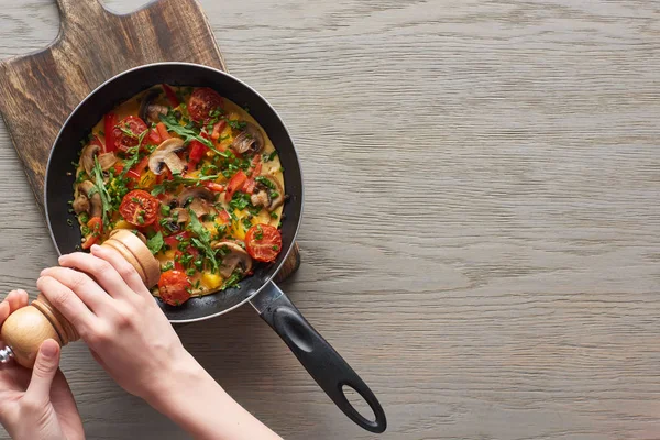 Cropped view of woman sprinkling pepper on tasty omelet in frying pan on wooden board — Stock Photo