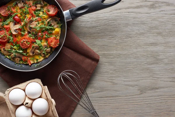 Top view of delicious omelet in frying pan with eggs and whisk on table — Stock Photo