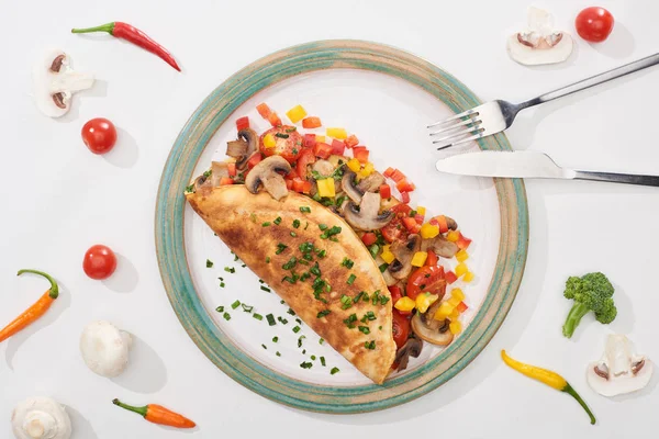 Vue du dessus de la plaque avec omelette enveloppée savoureuse avec légumes sur table blanche avec ingrédients, fourchette et couteau — Photo de stock