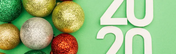 Top view of white 2020 numbers near multicolored Christmas baubles on green background, panoramic shot — Stock Photo