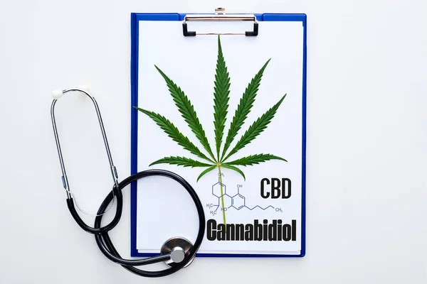 Vista superior da folha de cannabis medicinal na área de transferência com letras cbd perto do estetoscópio no fundo branco — Fotografia de Stock
