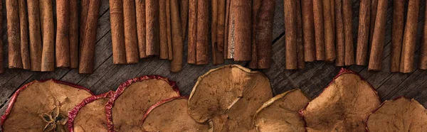 Panoramic shot of dried apple slices and cinnamon sticks on wooden background — Stock Photo