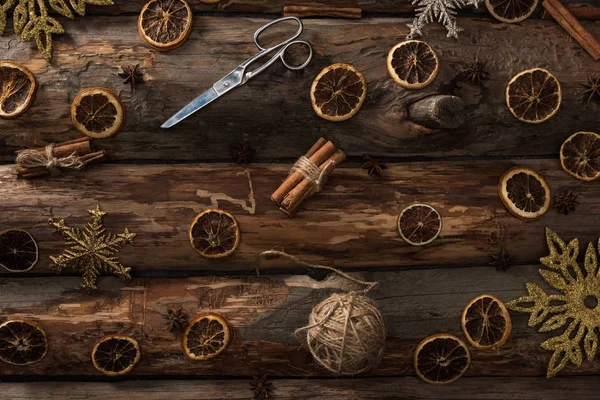 Top view of cinnamon sticks, anise, dried citrus slices, scissors, ball of thread and snowflakes on wooden background — Stock Photo