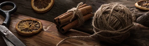 Close up view of cinnamon sticks, scissors, ball of thread on wooden background with dried citrus slices, panoramic shot — Stock Photo