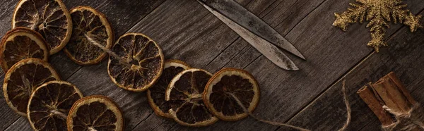 Top view of dried citrus slices on rope near snowflake, cinnamon and scissors on wooden surface, panoramic shot — Stock Photo